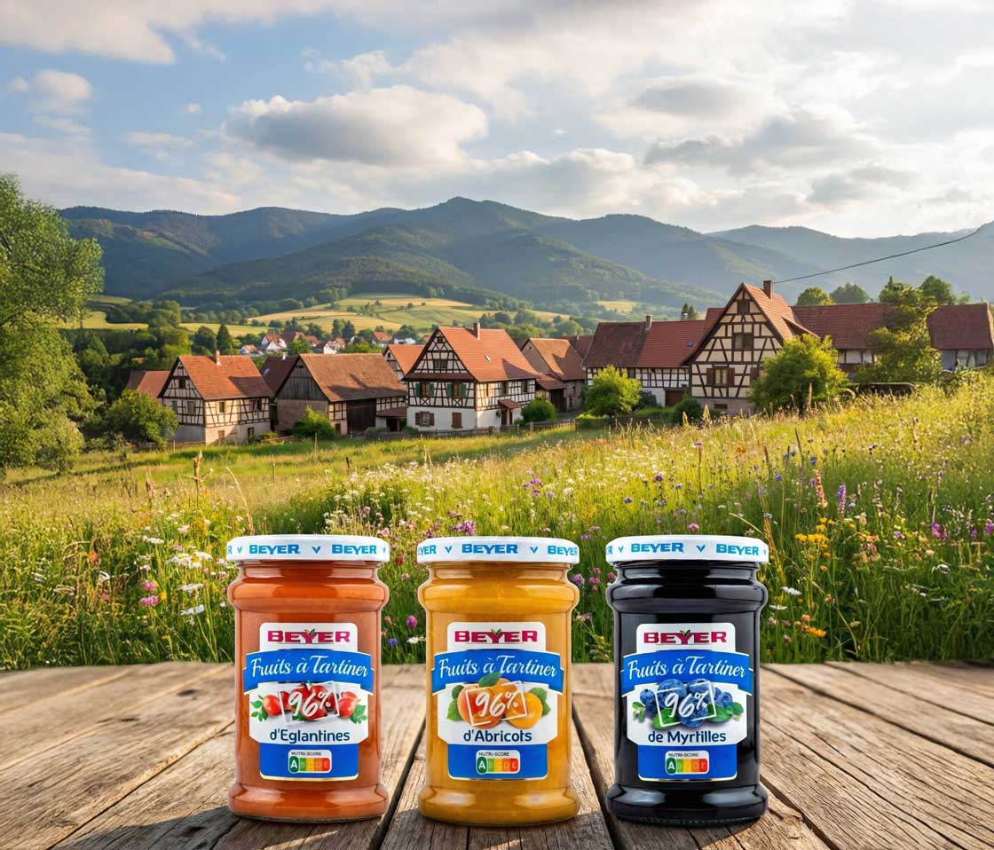 Deux pots de la gamme Beyer "Les 100%" (Cerises Noires et Rhubarbe) posés sur une table en bois rustique. L'arrière-plan montre un village alsacien pittoresque niché dans une vallée verdoyante au pied de montagnes escarpées.